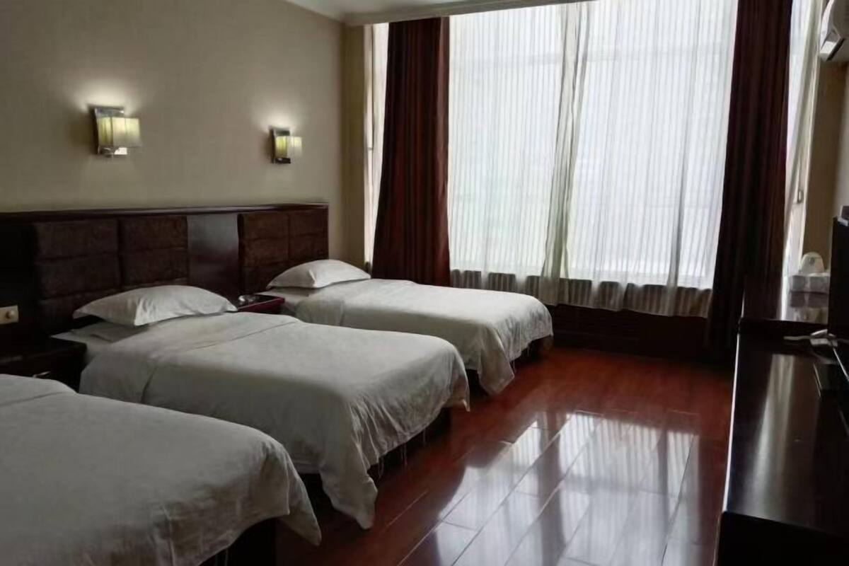 A hotel room with three beds, a wooden headboard, a large window with curtains, and wall-mounted lights.