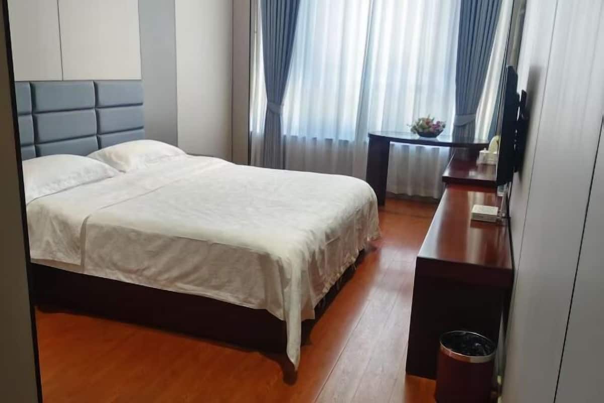 A hotel room with a double bed, a wooden floor, a desk with a chair, a TV, and a window with curtains.