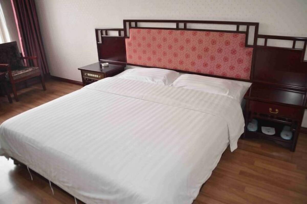 A neatly made bed with a wooden headboard, a bedside table with a drawer, and a small shelf with slippers.