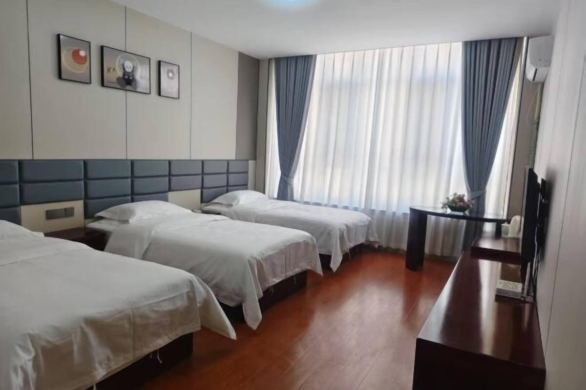 A hotel room with three beds, a wooden floor, a desk with a chair, and a large window with curtains.