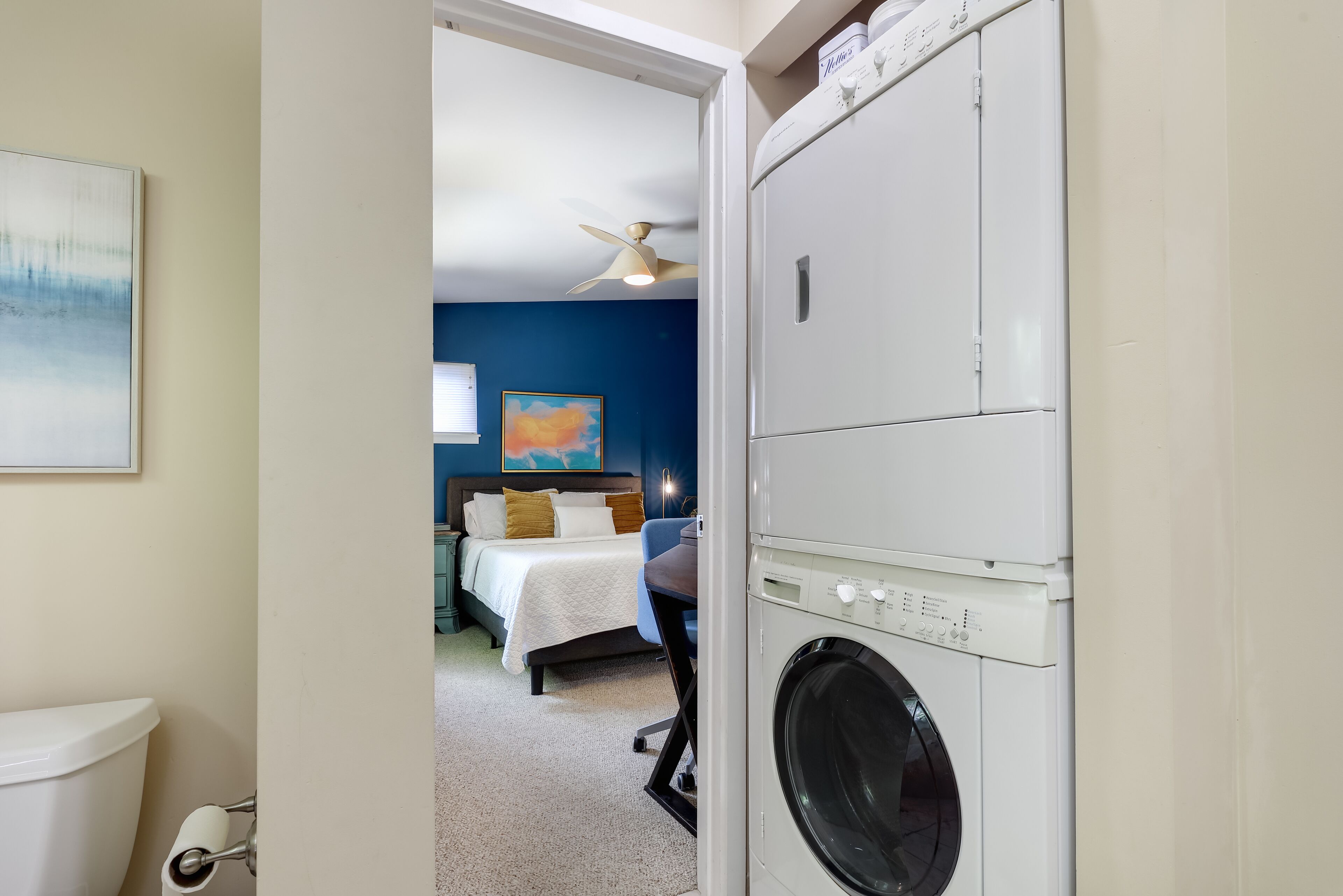 Ein Hotelzimmer mit Waschmaschine, einem Bett mit weißer Bettdecke, einem Deckenventilator und einem Gemälde an der Wand.
