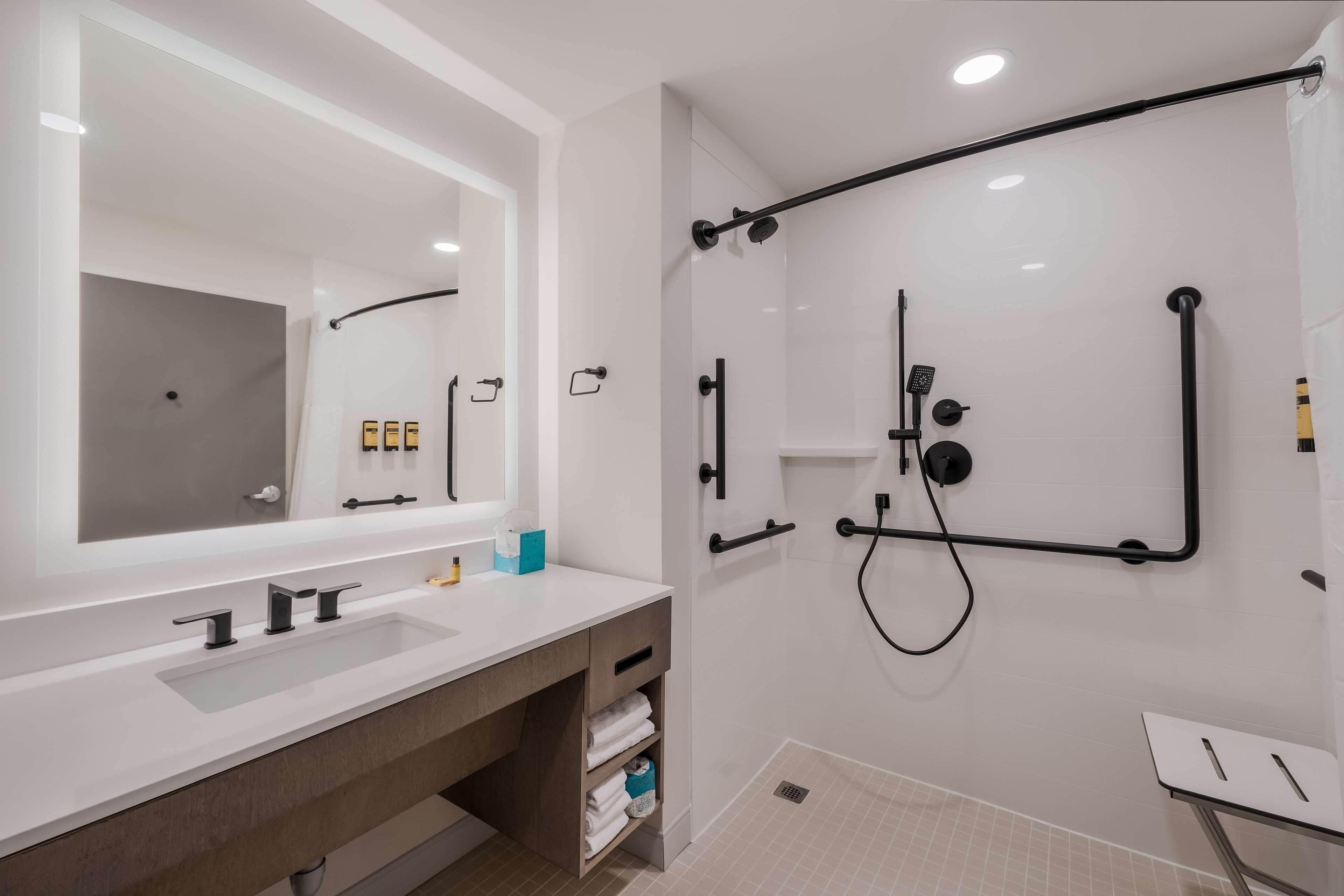 A modern bathroom with a large mirror, dual sinks, a shower with a black frame, and a small table.