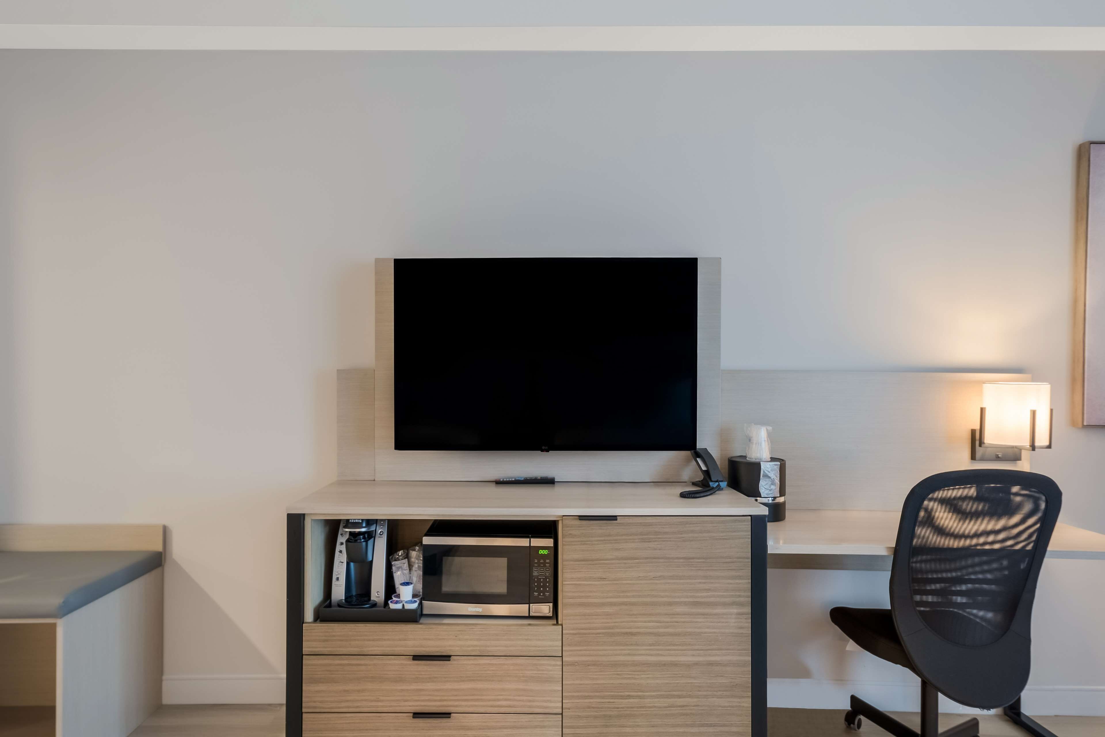 Et moderne hotelværelse med et fladskærms-TV monteret på et træskrivebord, en mikroovn og en sort kontorstol.