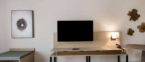 Quarto moderno com televisão de tela plana fixada em um painel de madeira, uma mesa de cabeceira, uma cadeira e uma pequena mesa redonda.