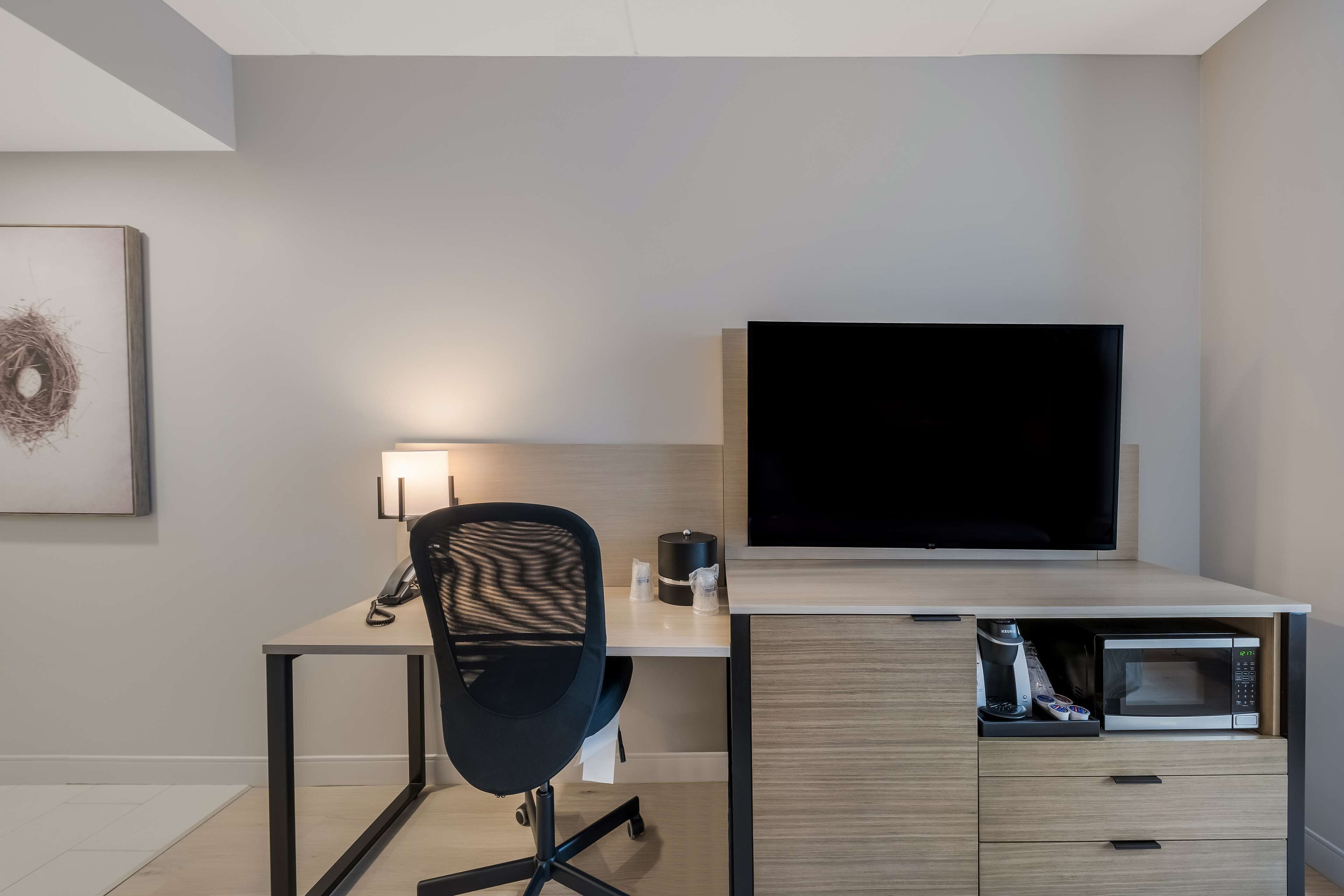 A modern office space with a desk, chair, flat-screen TV, lamp, and built-in microwave.