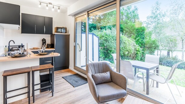 A modern kitchen with a dining area, a large sliding glass door leading to a garden, and a comfortable seating area with a chair and table. - Studio Aristide - Welkeys (Cabourg)