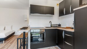 A modern kitchen with black cabinets, a built-in oven, and a dining area with a wooden table and chairs. - Studio Aristide - Welkeys (Cabourg)