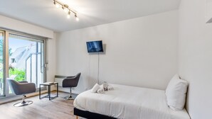 A modern bedroom with a large bed, a TV mounted on the wall, and a chair by the window. - Studio Aristide - Welkeys (Cabourg)