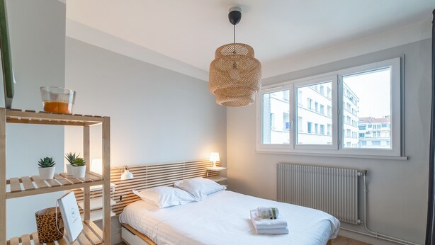 A modern bedroom with a wooden headboard, a large window with a view of buildings, a hanging rattan light fixture, and a bedside table with a lamp and a plant. - Appartement de la Bannière - Welkeys (Lyon)