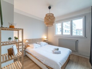 A modern bedroom with a wooden headboard, a large window with a view of buildings, a hanging rattan light fixture, and a bedside table with a lamp and a plant. - Appartement de la Bannière - Welkeys (Lyon)