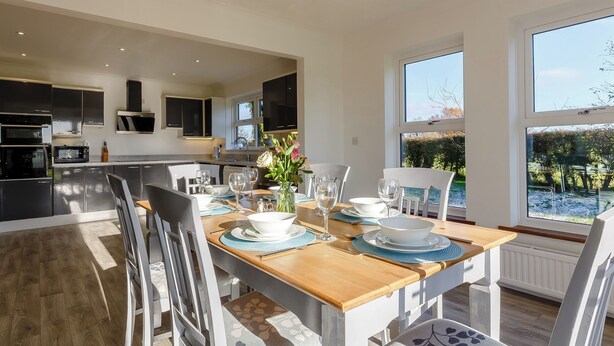 A modern kitchen with built-in appliances, a dining area with a wooden table and chairs, and large windows offering a view of the outdoors. - Lie back and count the stars whilst lazing in the hot tub. (Gissing, near Diss)