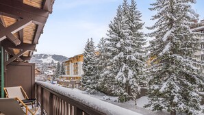 A snow-covered balcony with a wooden railing, a chair, and a view of snow-laden trees and buildings in the background.