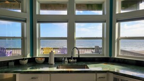 Une cuisine dotée d’une grande fenêtre donnant sur une plage, d’un évier et d’un plan de travail sur lequel se trouvent un saladier et des bouteilles.