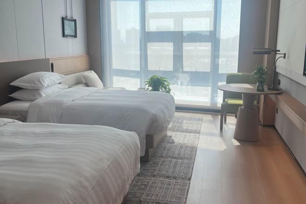 A hotel room with two beds, a small table, a chair, and a view of the city through large windows.