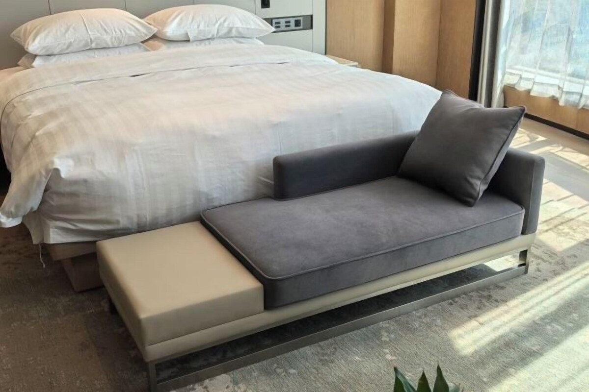 A modern bedroom with a large bed, a grey sofa, and a small bench.