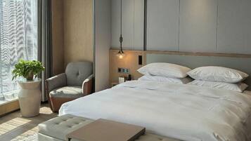 A modern hotel room with a large bed, a seating area, a potted plant, and a view through the window.