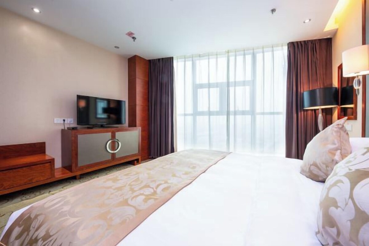 A hotel room with a bed, a television on a wooden cabinet, a large window with curtains, and a bedside lamp.