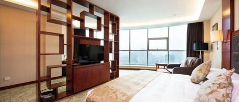 A hotel room with a large bed, a TV, a seating area, and a view of the city.