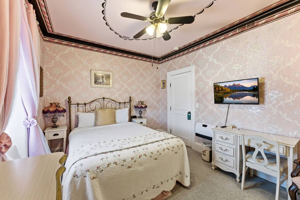 A bedroom with a bed, a desk, a ceiling fan, a TV, and a door.