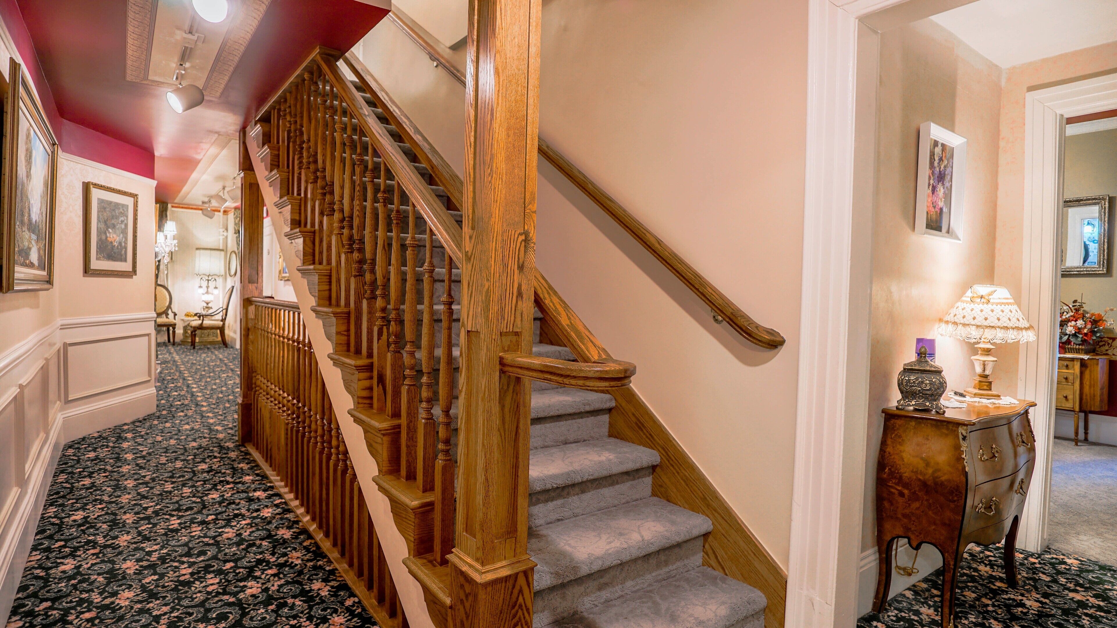 A grand staircase with wooden balusters and carpeted steps, leading to a landing with a lamp and framed artwork.