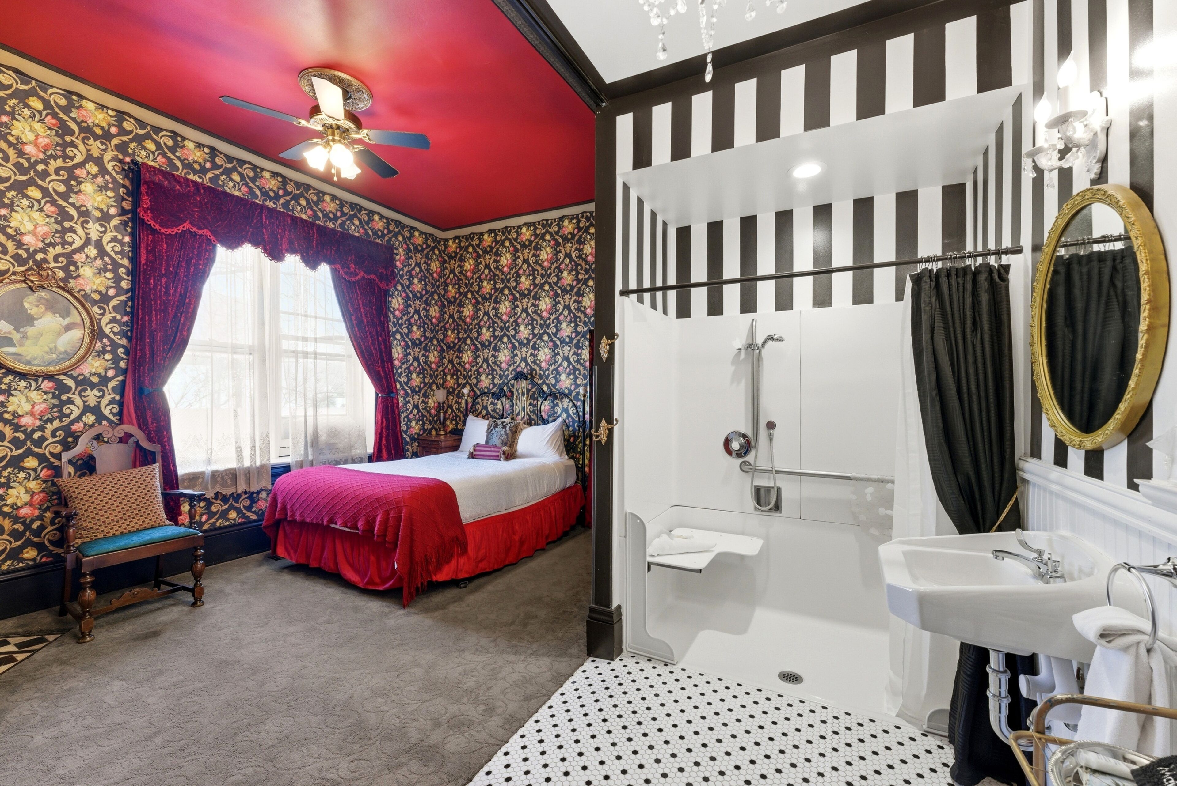 A bedroom with a bed, a chair, a window with curtains, a shower area, and a bathroom with a sink and mirror.