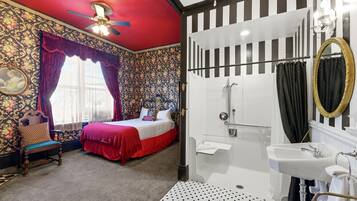 A bedroom with a bed, a chair, a window with curtains, a shower area, and a bathroom with a sink and mirror.