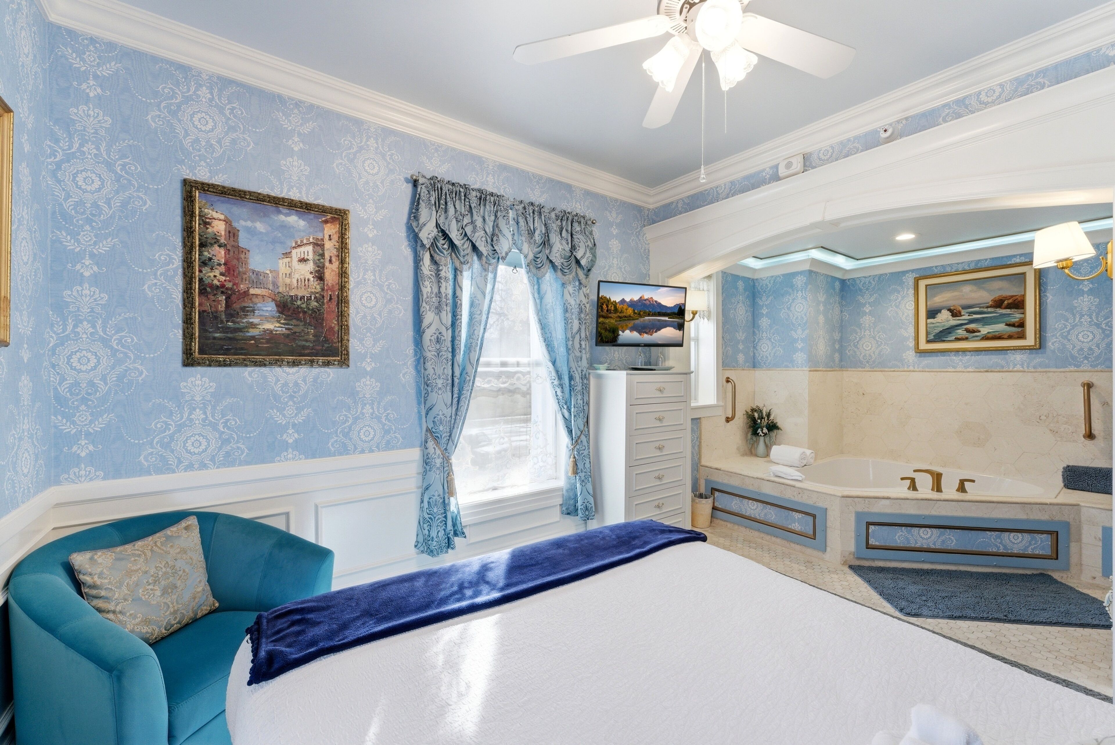 A bedroom with a bed, a blue chair, a vanity with a bathtub, and a ceiling fan.