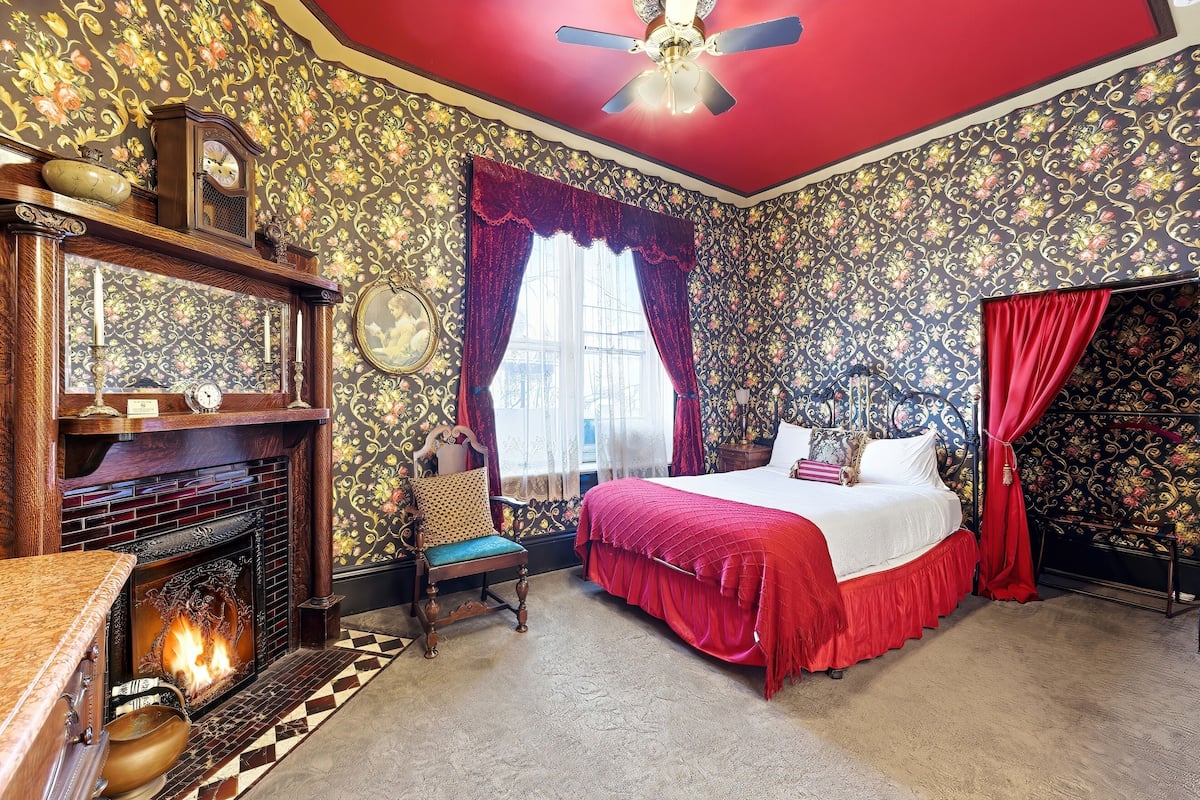 A room with a fireplace, a bed with red curtains, a clock, and patterned wallpaper.