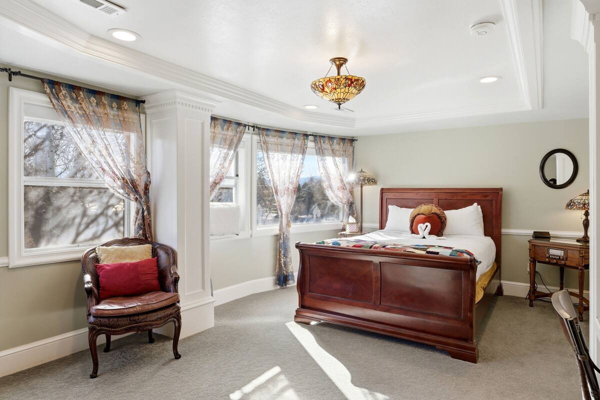 A bedroom with a large bed, a chair, a nightstand, a mirror, and a window with curtains.