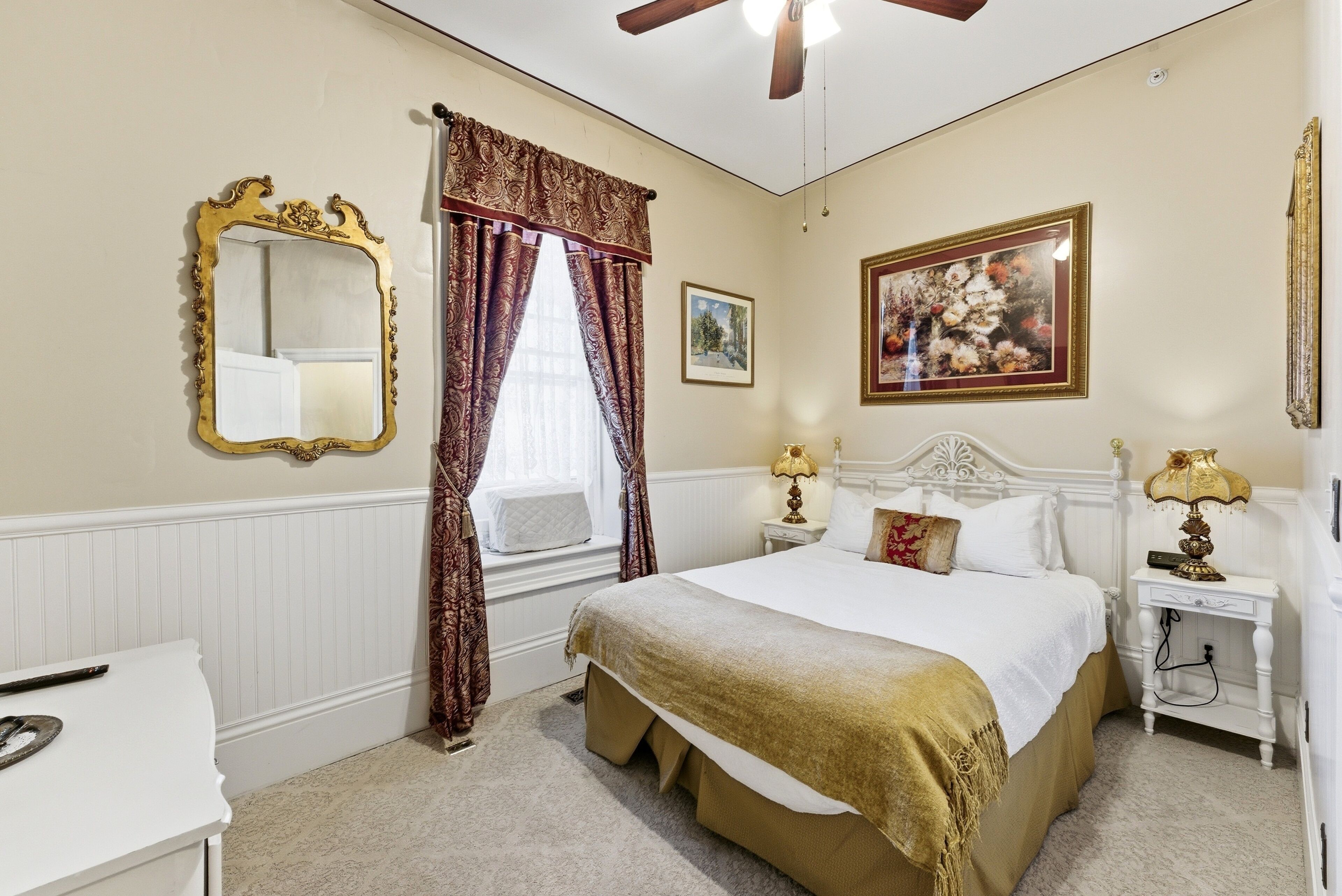A bedroom with a bed, a nightstand, a mirror, a window with curtains, and a ceiling fan.