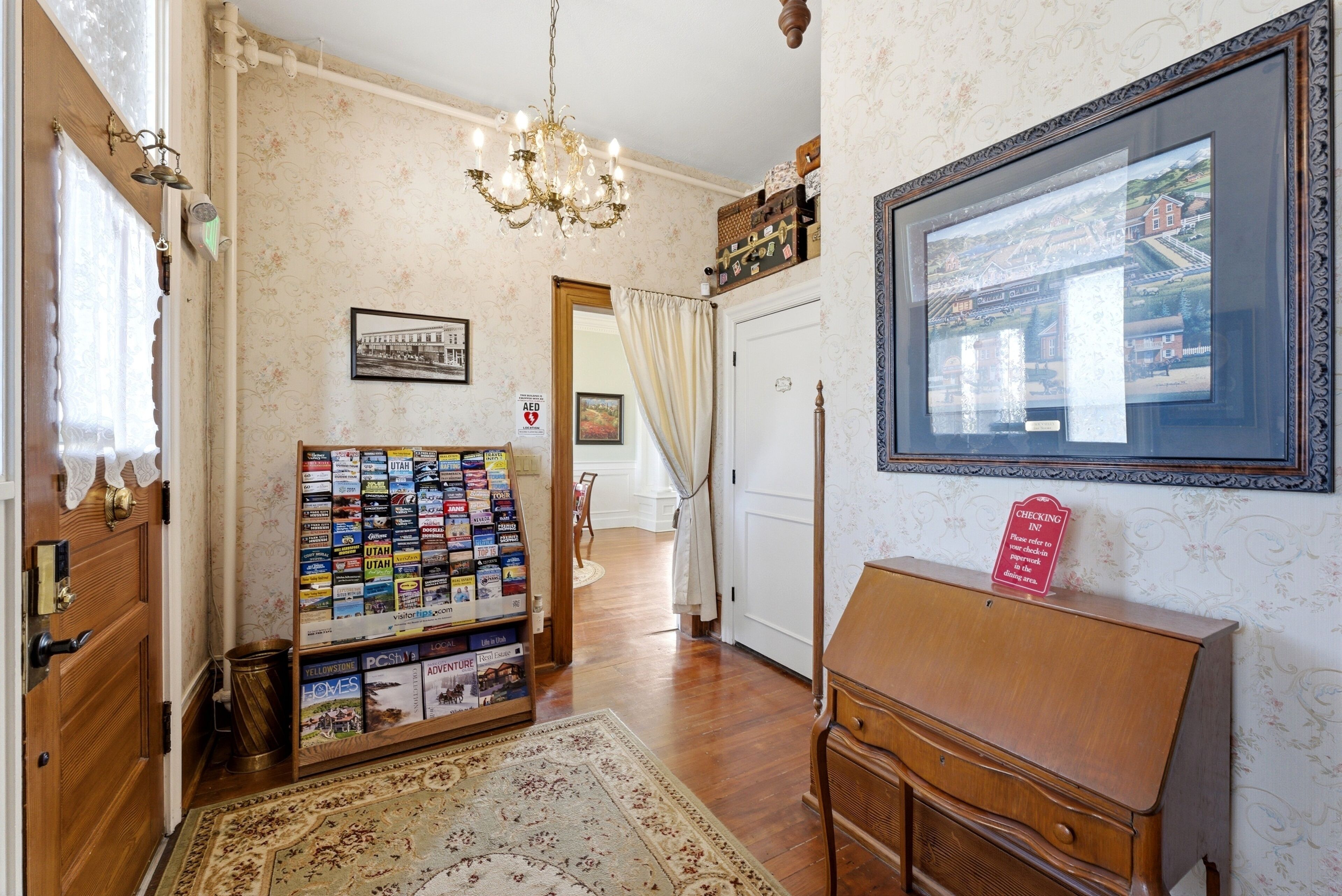 A hallway with a wooden floor, a chandelier, a framed picture on the wall, a check-in sign, and a magazine rack.