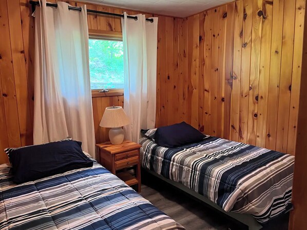 A room with two beds, wooden walls, a window with curtains, a nightstand with a lamp, and a view of greenery outside.