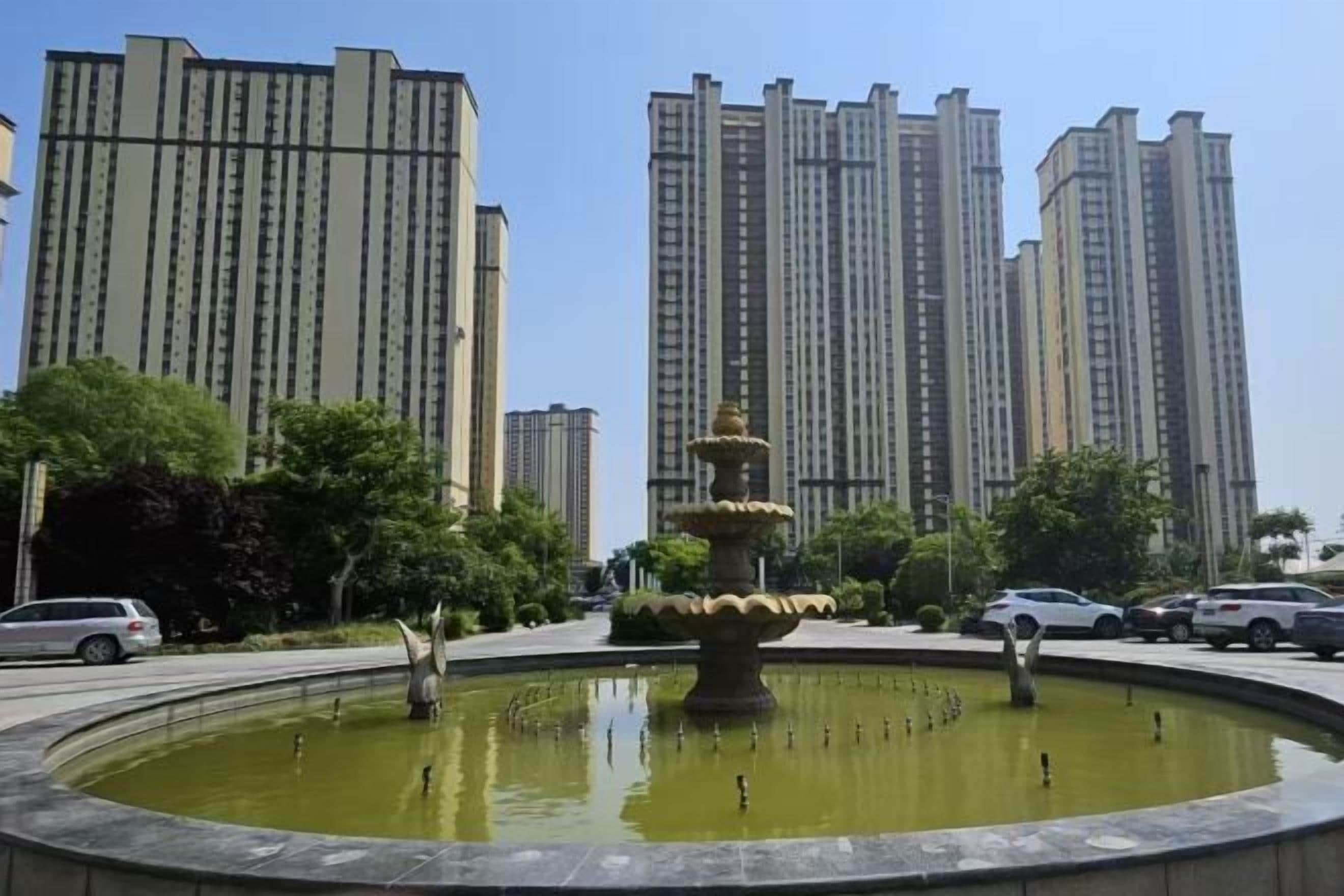 A circular fountain with tiered water features surrounded by a paved area, with tall residential buildings in the background.
