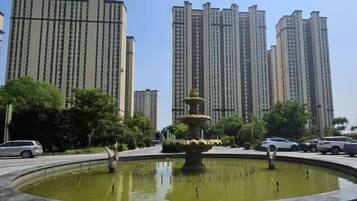A circular fountain with tiered water features surrounded by a paved area, with tall residential buildings in the background.