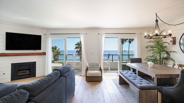 A modern living room with a large TV, a fireplace, a sofa, a dining table with chairs, and a balcony with a view of the ocean. - Beachfront Hideaway in Santa Barbara - 2 bedrooms, pool, spa, across from beach, gorgeous! (Santa Barbara)