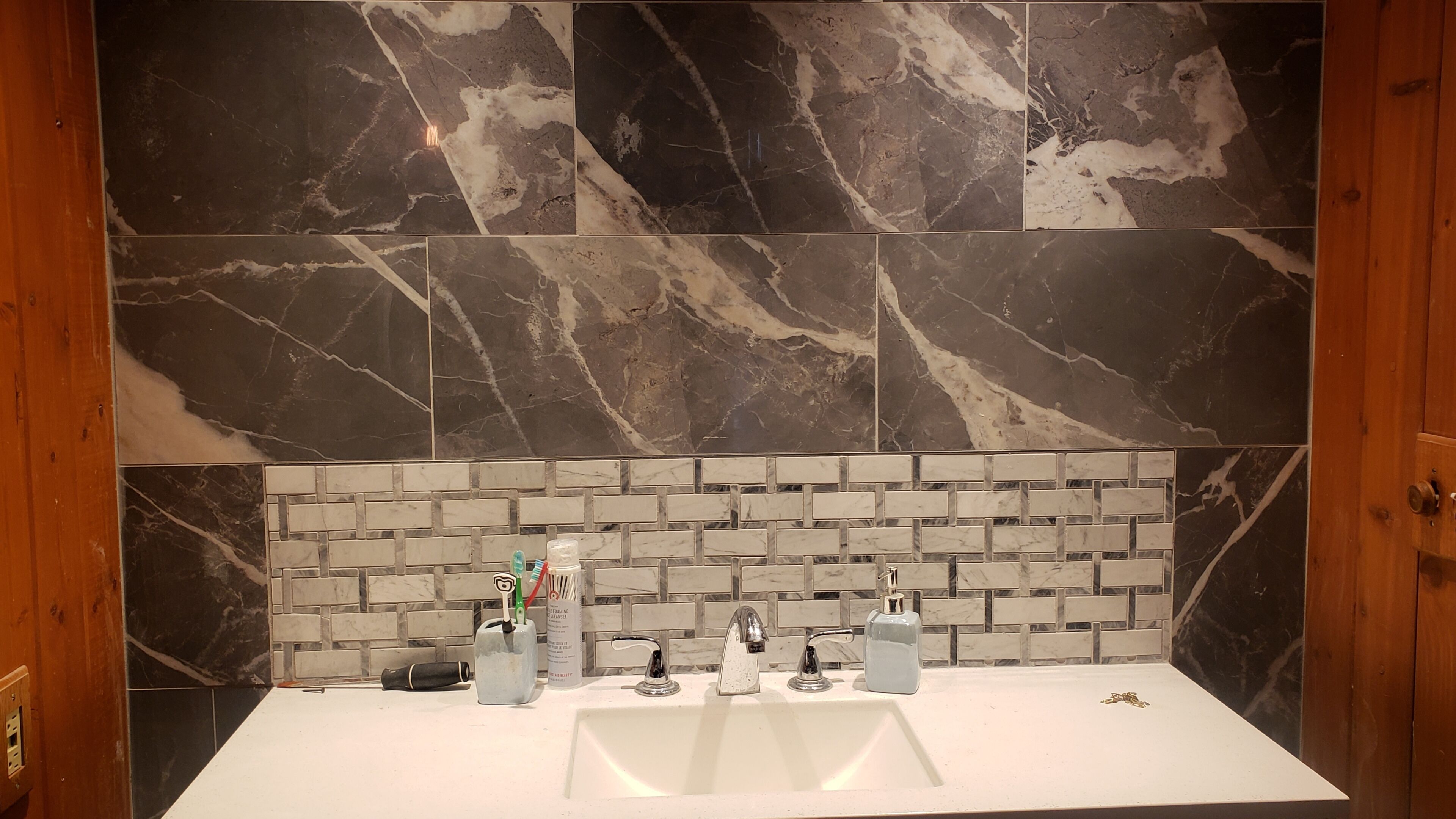 Ein Badezimmer mit einem weißen Waschbecken, zwei Wasserhähnen und einer Seifenspender. Die Wand hinter dem Waschbecken zeigt ein schwarz-weiß Marmormuster.