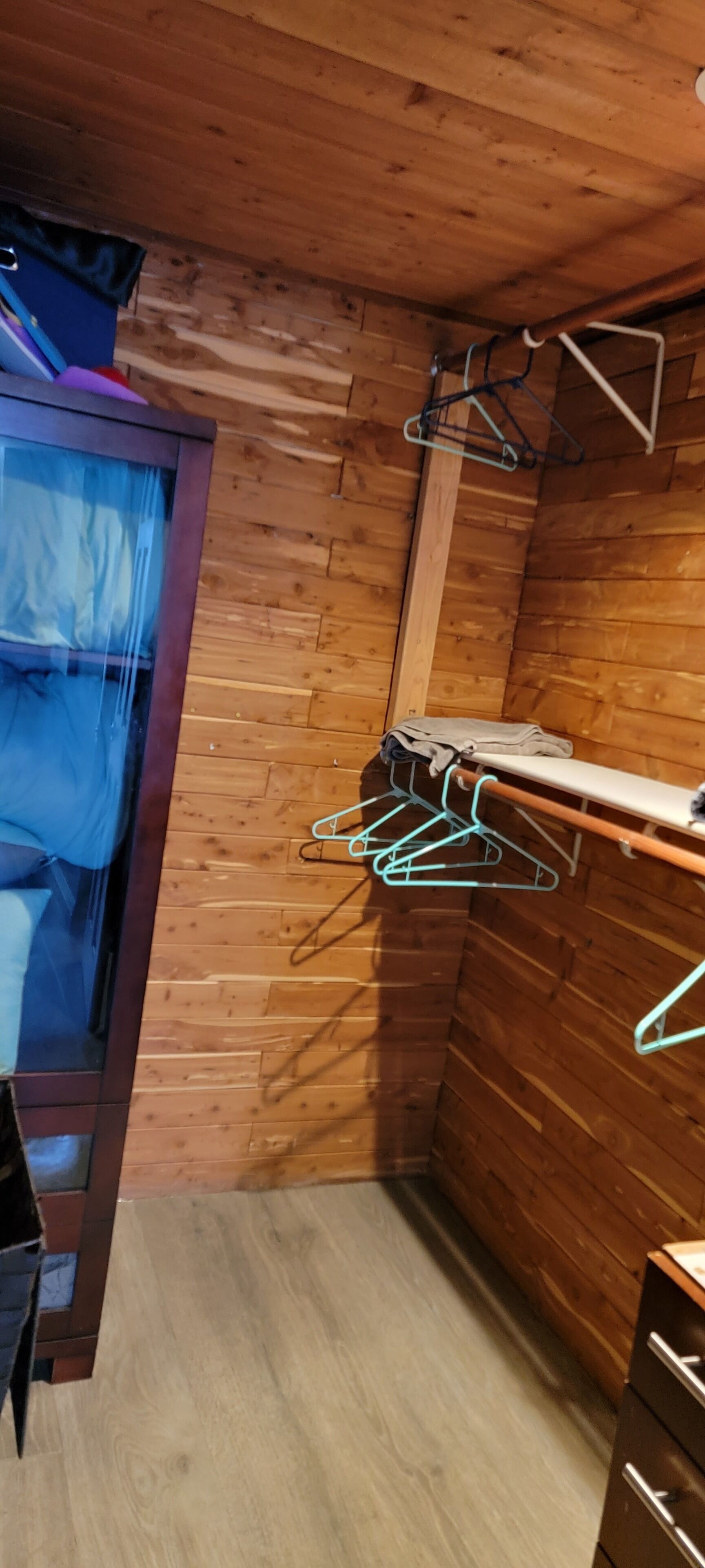 Rincón de una habitación con paredes y suelo de madera, un tendedero de metal con ropa y un estante pequeño.
