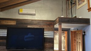 Ein Zimmer mit Holzboden, einem an der Wand befestigten Flachbildfernseher, einem Deckenventilator und einer Tür zu einem weiteren Raum.