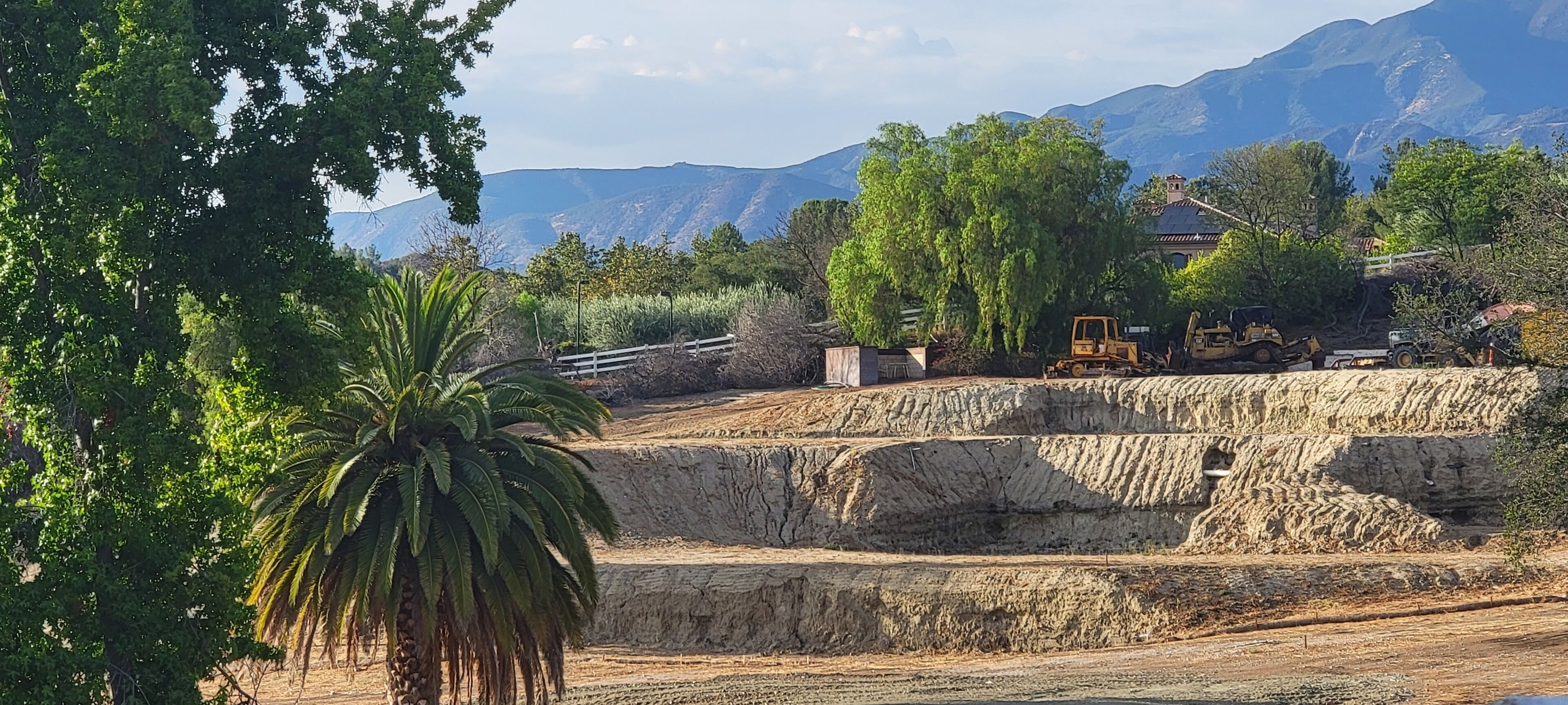 Un chantier de construction avec une excavatrice jaune, une clôture blanche et une montagne en arrière-plan.