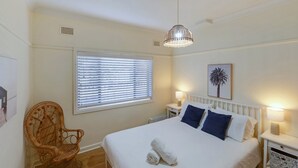 A bedroom with a bed, two bedside tables with lamps, a window with blinds, a chair, and a wall-mounted light fixture.