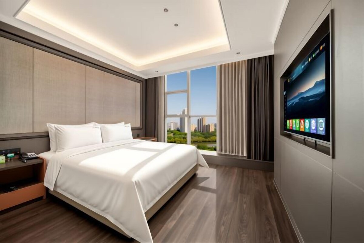 A modern hotel room with a large bed, a TV mounted on the wall, and a window offering a city view.
