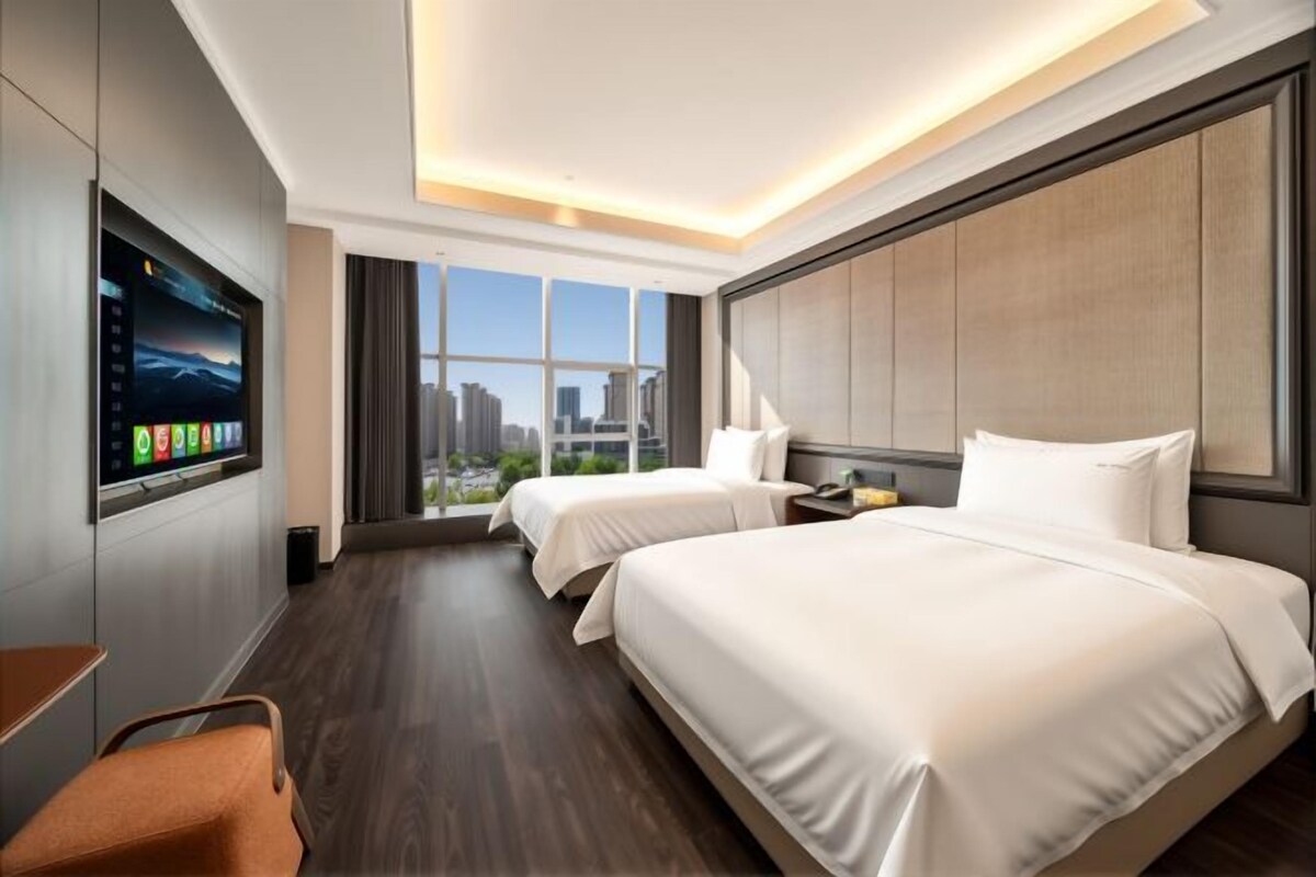 A hotel room with two beds, a TV, a desk, and a window with a city view.