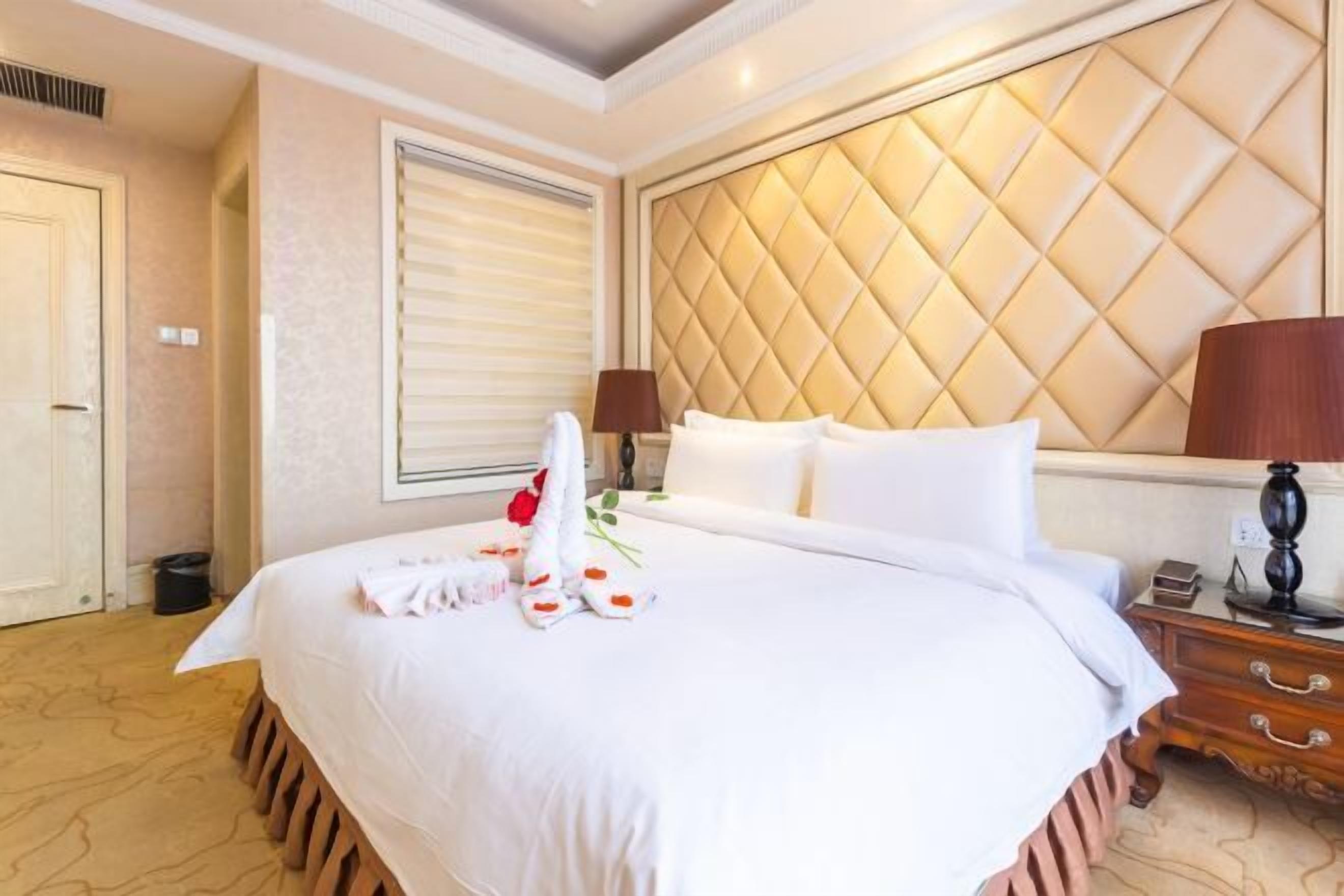 Une chambre d’hôtel avec un grand lit, une tête de lit ornée d’un motif en losanges, une table de chevet avec une lampe et une fenêtre avec des stores.