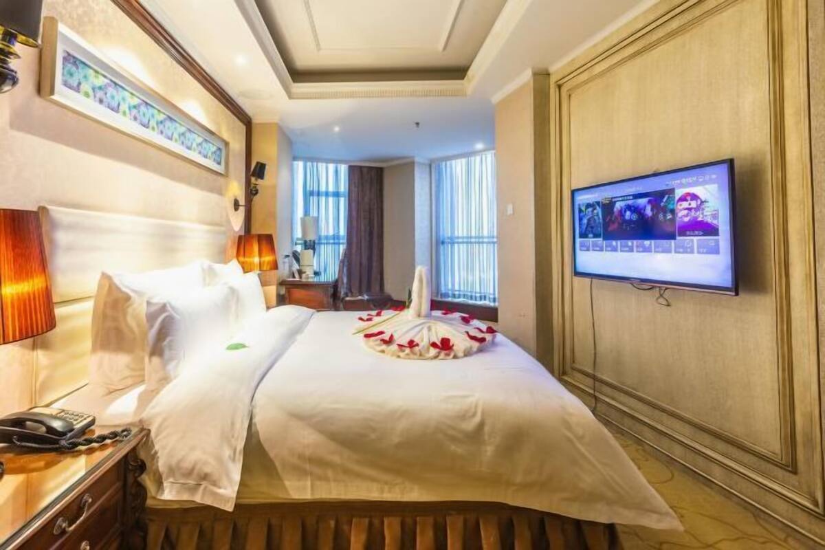 Ett hotellrum med en säng, en tv, ett skrivbord med en telefon och ett fönster med gardiner.