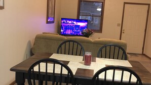 Un área de comedor con una mesa y cuatro sillas, un televisor mostrando un paisaje urbano, y una ventana con persianas.