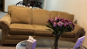 Una sala de estar con un sofá color beige, una mesa de centro de vidrio, un jarrón con flores, una caja de pañuelos y un cuadro en la pared.