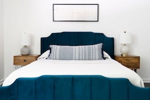 Una cama bien hecha con cabecera color turquesa y ropa de cama blanca, flanqueada por dos mesitas de noche con lámparas y un cuadro enmarcado encima.