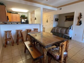 A spacious room with a wooden dining table, four stools, a bench, a bed with a patterned bedspread, and a kitchen area with wooden cabinets and a refrigerator. - Bear's Den • Walk to attractions • Mountain View Condo • Virtual Checkin (Pigeon Forge)