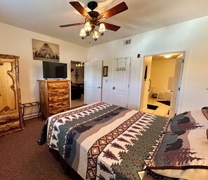 A bedroom with a large bed, a ceiling fan, a wooden dresser, a television, and a door leading to another room. - Bear's Den • Walk to attractions • Mountain View Condo • Virtual Checkin (Pigeon Forge)
