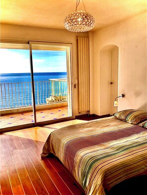 A bedroom with a large bed, a view of the ocean through a sliding door, and a chandelier. - The big VENTILATED blue (Corniche Roucas Blanc beach) (Marseille)